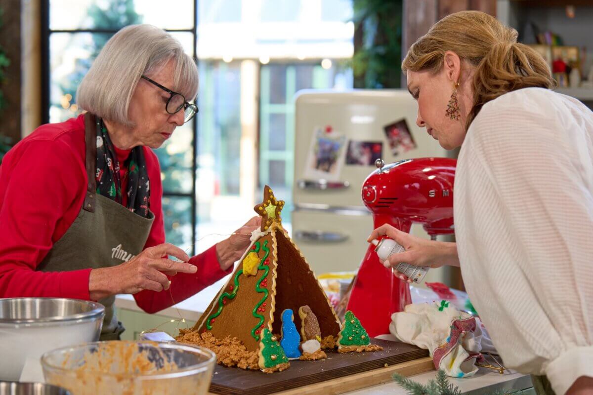 Meet Sally McKay, the Founder of Sally's Confections - 6 Sally McKay and Amy Wicker decorates a gingerbread house shaped like a tent with frosting and candy, working together in a bright kitchen.