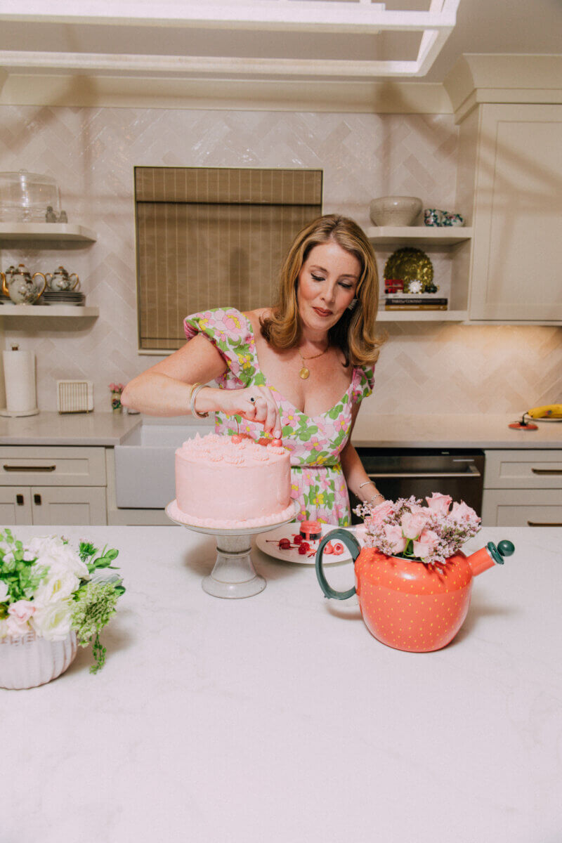 Meet Sally McKay, the Founder of Sally's Confections - 2 Sally McKay decorates a pink cake on a stand in a kitchen with light-colored cabinets and floral arrangements.