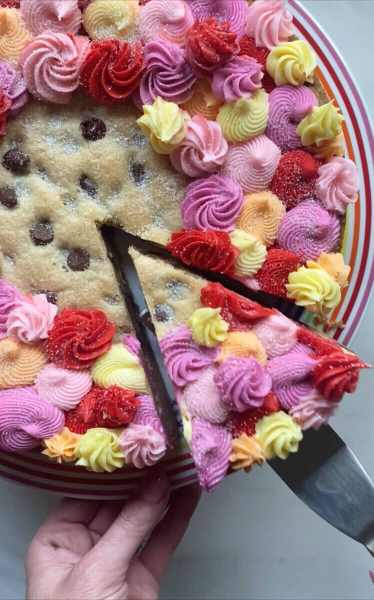 Meet Sally McKay, the Founder of Sally's Confections - 3 A slice is being cut from a large cookie cake baked with love, decorated with colorful swirled icing in pink, red, yellow, and purple on a striped plate.