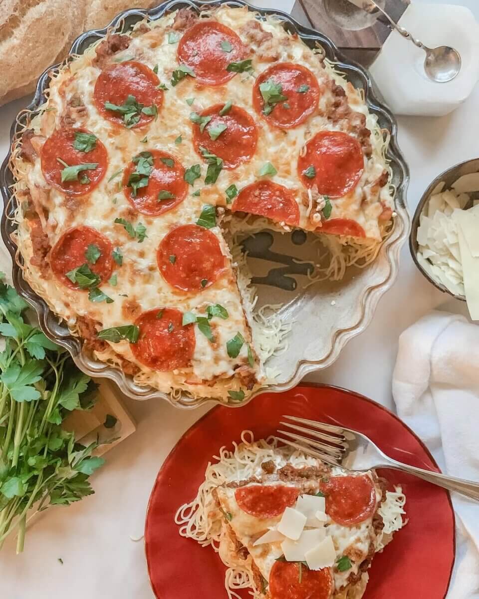 Meet Sally McKay, the Founder of Sally's Confections - 8 A pepperoni and cheese pizza pie, baked with love, has a slice removed and is served on a red plate with a fork. Fresh herbs, shredded cheese, and bread are nearby.