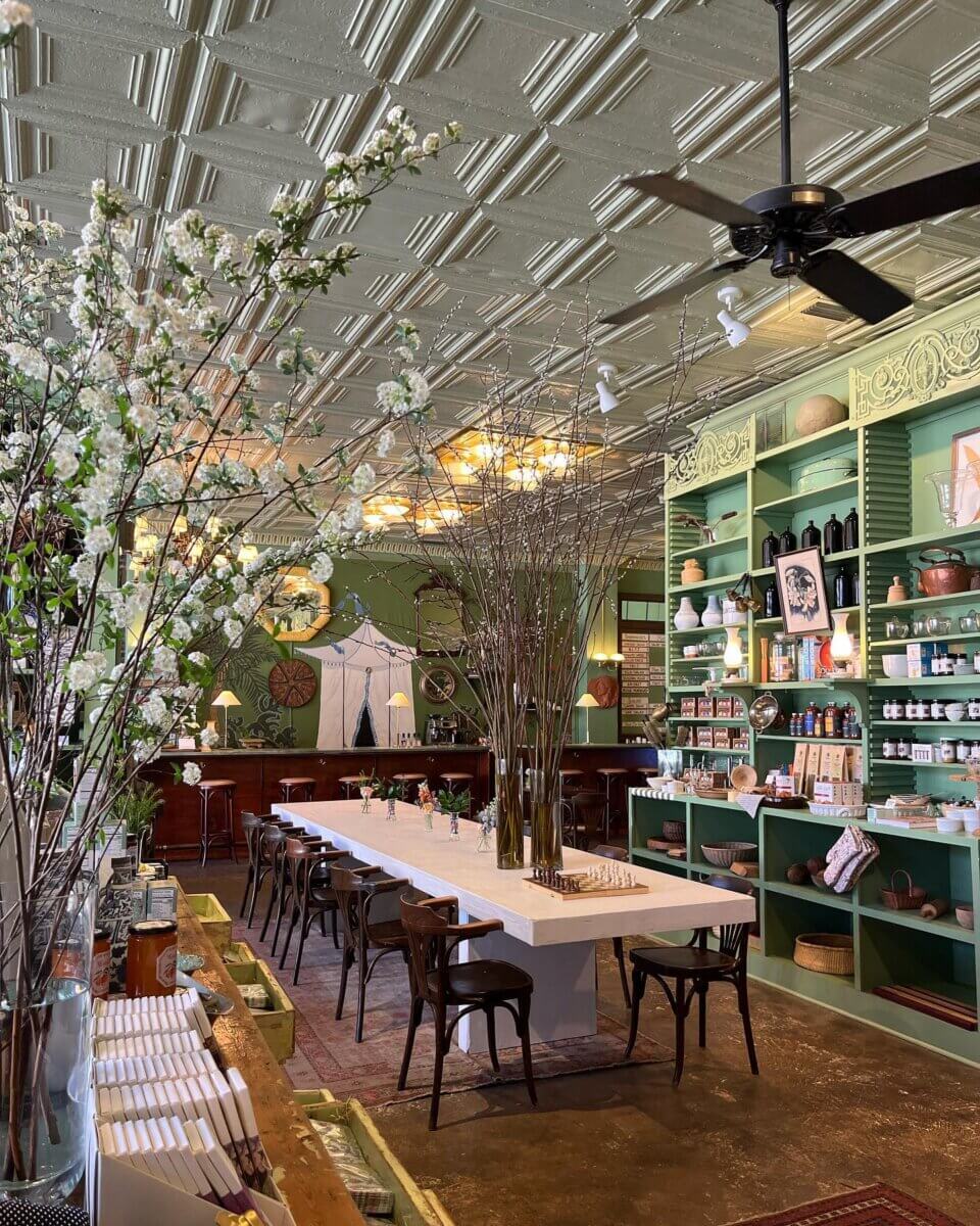 Your Guide to BHAM's Forest Park Neighborhood - 3 A long communal table with wooden chairs is set in a cozy Forest Park shop in Birmingham, featuring green walls, high shelves of goods, tall vases of branches, and a decorative ceiling.