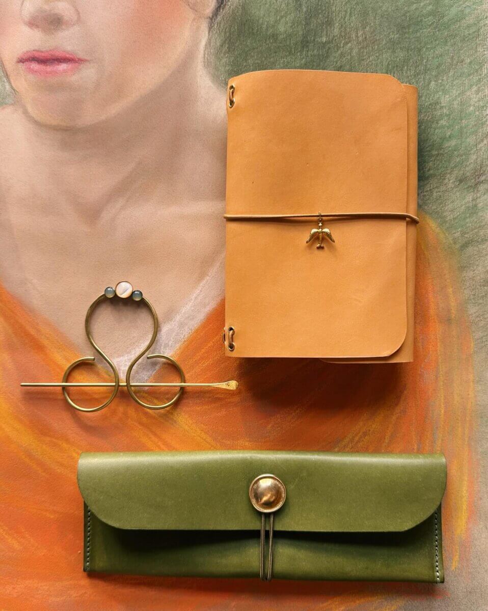 Your Guide to BHAM's Forest Park Neighborhood - 4 A brown leather notebook, a green leather case with a button, and a gold hairpin lay on a pastel portrait background of a person in orange.