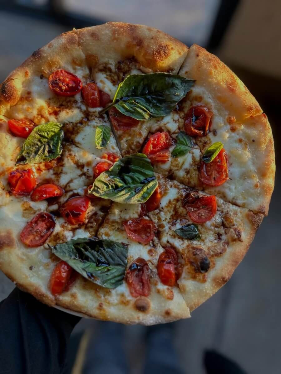 Your Guide to BHAM's Forest Park Neighborhood - 2 A hand holds a pizza topped with melted cheese, cherry tomatoes, and fresh basil leaves, sliced into six pieces—perfect for sharing in Forest Park, BHAM’s favorite foodie neighborhood.