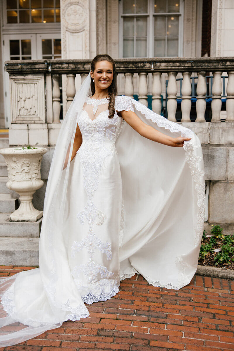 A woman in a white wedding dress with lace details and a long train stands outside the Linn-Henley Research Library on a brick path, holding part of her dress and smiling, creating a true storybook wedding moment.