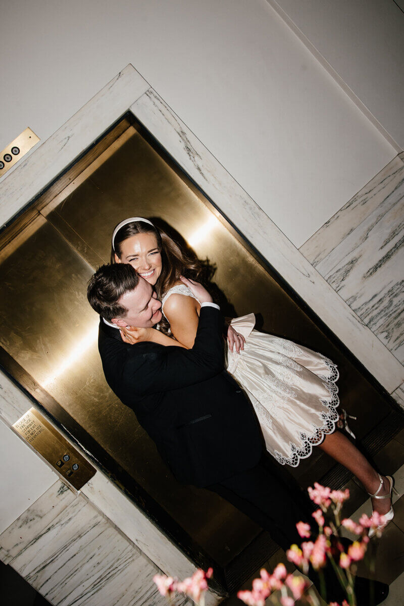 A man and woman dressed formally embrace and laugh in front of a closed elevator with marble trim at the stunning wedding venue, Linn-Henley Research Library. Pink flowers are visible in the foreground, adding to their storybook wedding moment.