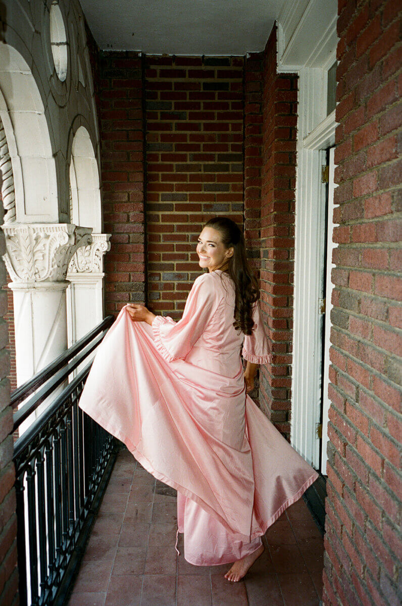 A woman in a long pink dress stands barefoot on a balcony with brick walls at the Linn-Henley Research Library, looking back over her shoulder and holding her dress—capturing a storybook wedding moment.