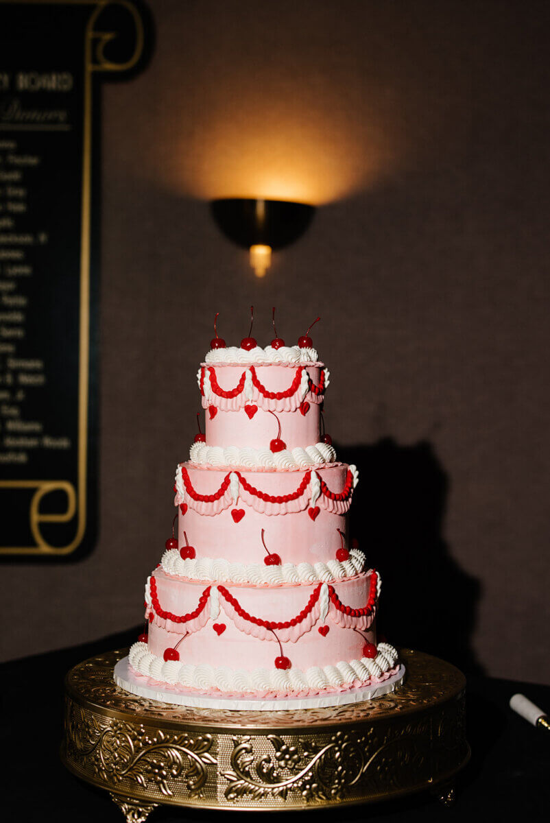 A three-tier pink cake decorated with red hearts, cherries, and white icing sits on a gold cake stand against a dark background—perfect for a storybook wedding at the Linn-Henley Research Library.