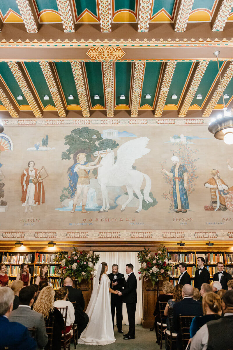 A bride and groom stand at the altar with an officiant in the Linn-Henley Research Library, beneath a large Pegasus mural—surrounded by bookshelves and guests, this storybook wedding unfolds in a stunning wedding venue.