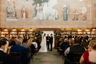 A storybook wedding unfolds at the Linn-Henley Research Library, with the couple at the altar, surrounded by their wedding party and guests seated beneath a large mural in this stunning wedding venue. Photo by Alyssa Rose Photography