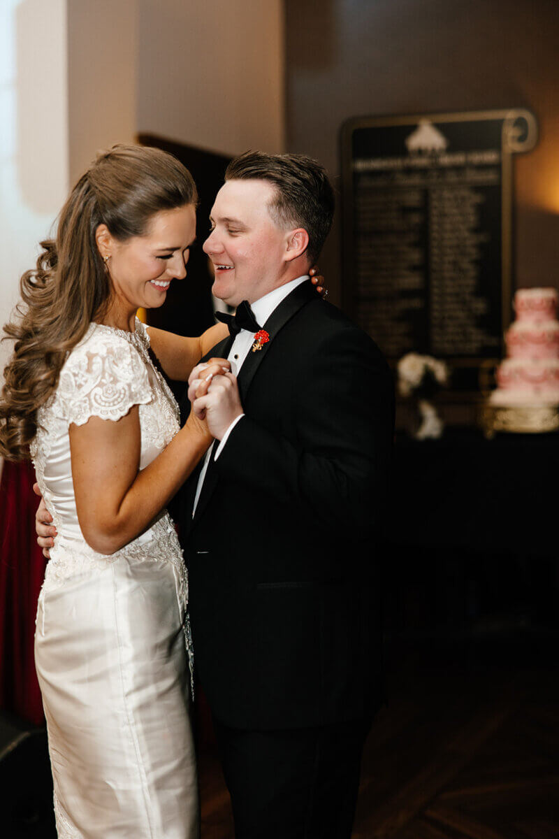 A bride and groom in formal attire share a dance, smiling at each other in the warmly lit Linn-Henley Research Library—a stunning wedding venue—with a cake visible in the background.