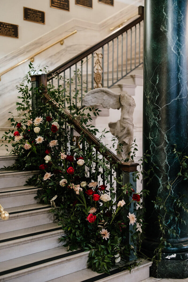 A marble staircase at the Linn-Henley Research Library, adorned with greenery and red and white flowers, creates a storybook wedding scene featuring a classical winged statue and plaques on the wall in the background.