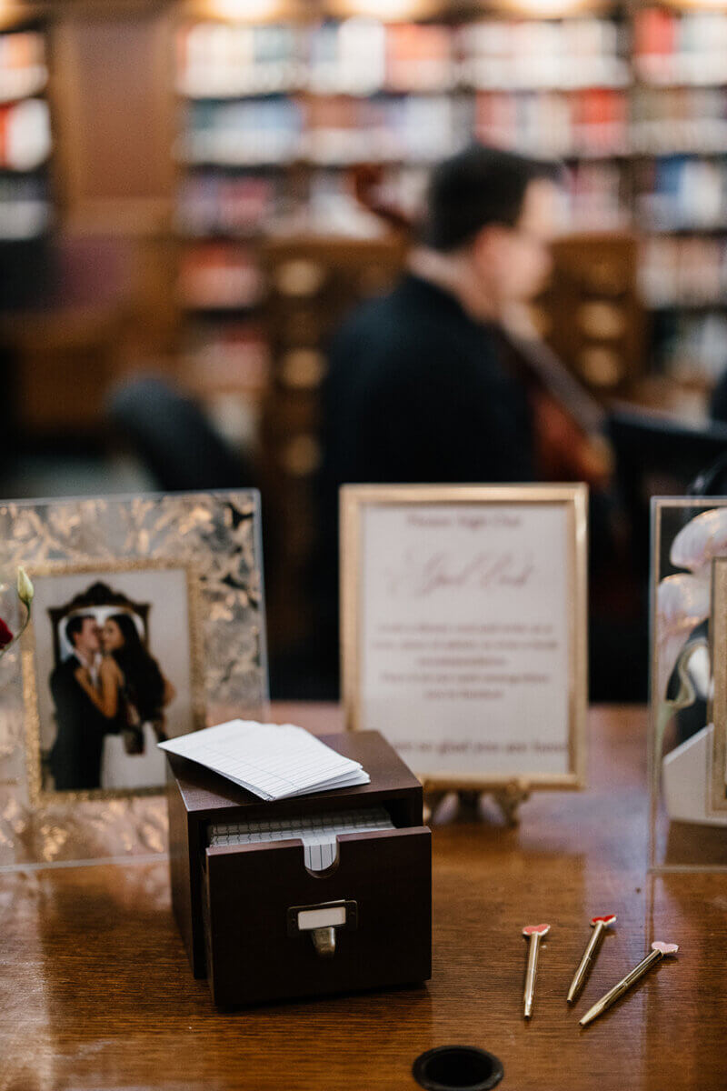 A wooden box with notecards and gold pens sits on a table at the stunning wedding venue, Linn-Henley Research Library; framed photos, a sign, blurred bookshelves, and a guest complete this storybook wedding scene.