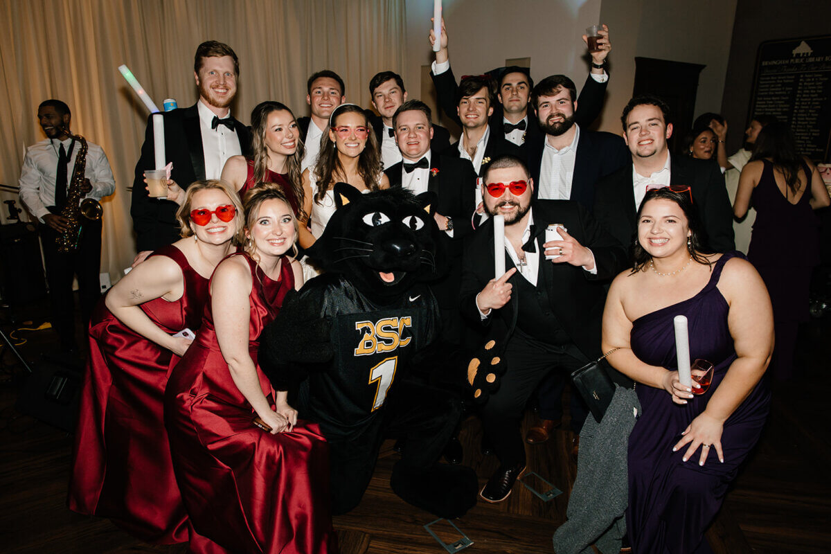 A group of people in formal attire pose with a panther mascot wearing a "BSU" jersey at the Linn-Henley Research Library, some holding glow sticks and smiling at the camera as if celebrating a storybook wedding.