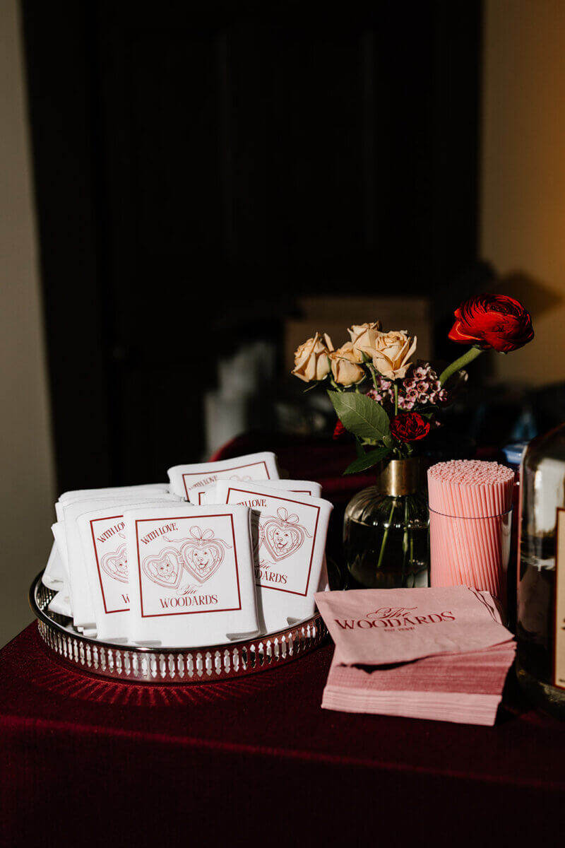 A silver tray holds custom playing cards with heart illustrations, next to pink napkins, a vase with flowers, and pink straws on a burgundy tablecloth—perfect details for a storybook wedding at the Linn-Henley Research Library.