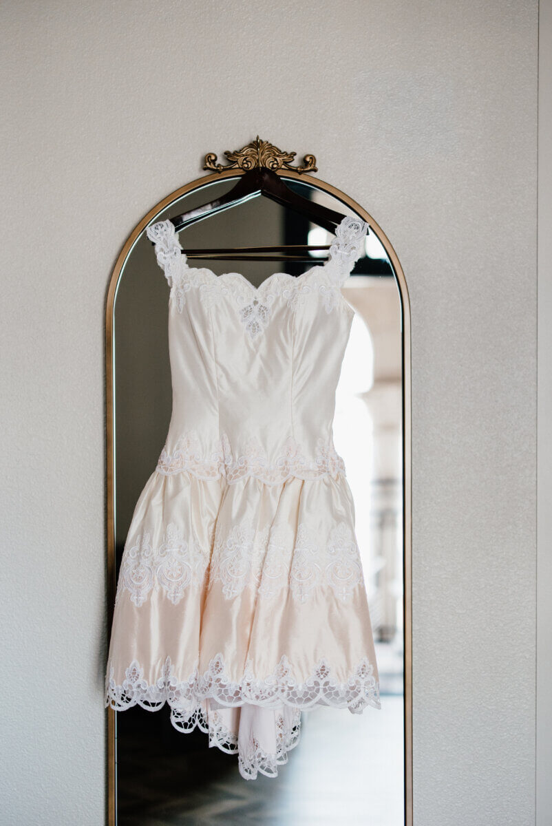 A white lace-trimmed dress hangs on a hanger in front of a tall, ornate mirror, evoking the charm of a storybook wedding against a light-colored wall.