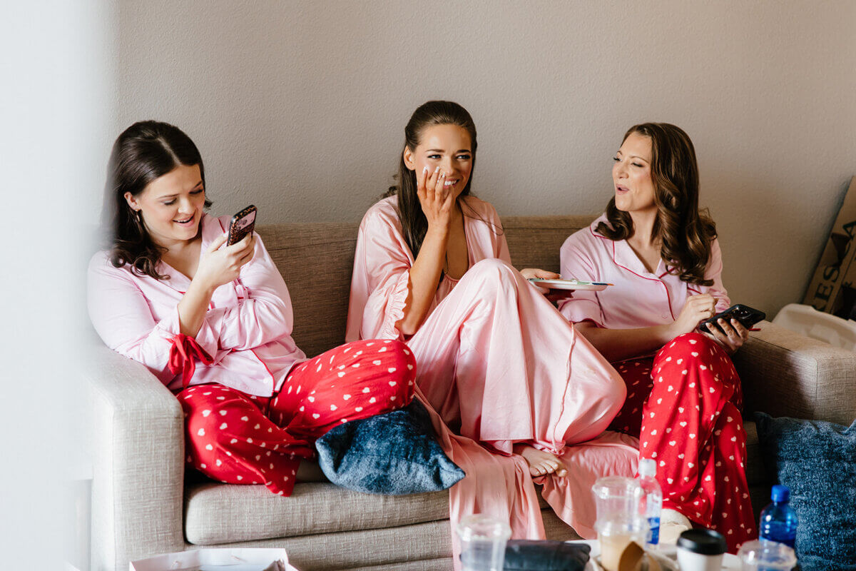 Three women in matching pink and red pajamas sit on a couch, laughing and looking at their phones and a tablet as they excitedly plan a stunning wedding at the Linn-Henley Research Library.