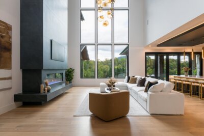 Modern living room with high ceilings, large windows, a white sectional sofa, a contemporary fireplace, and a view of greenery outside—a stunning example of real life design by C3 Studio.