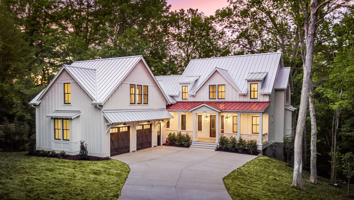 More Than a Floor Plan: C3 Studio Designs for Real Life - 3 A two-story modern farmhouse with a metal roof, large windows, a double garage, and a paved driveway—this real life design by C3 Studio is surrounded by trees at dusk.