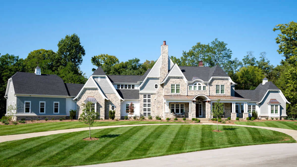 More Than a Floor Plan: C3 Studio Designs for Real Life - 2 Large stone and wood house with multiple gables, a chimney, and a circular driveway—showcases real life design by C3 Studio—set on a manicured lawn with trees in the background under a clear blue sky.