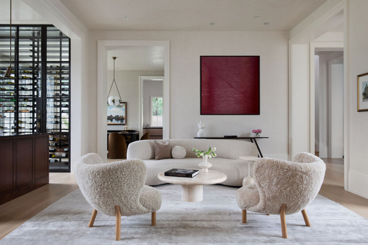 This Dallas Home Masters the Art of Quiet Luxury - 2 Modern living room with two cream armchairs, a matching sofa, round coffee table, wall art, and open view into adjacent rooms. Wine rack is visible on the left side.