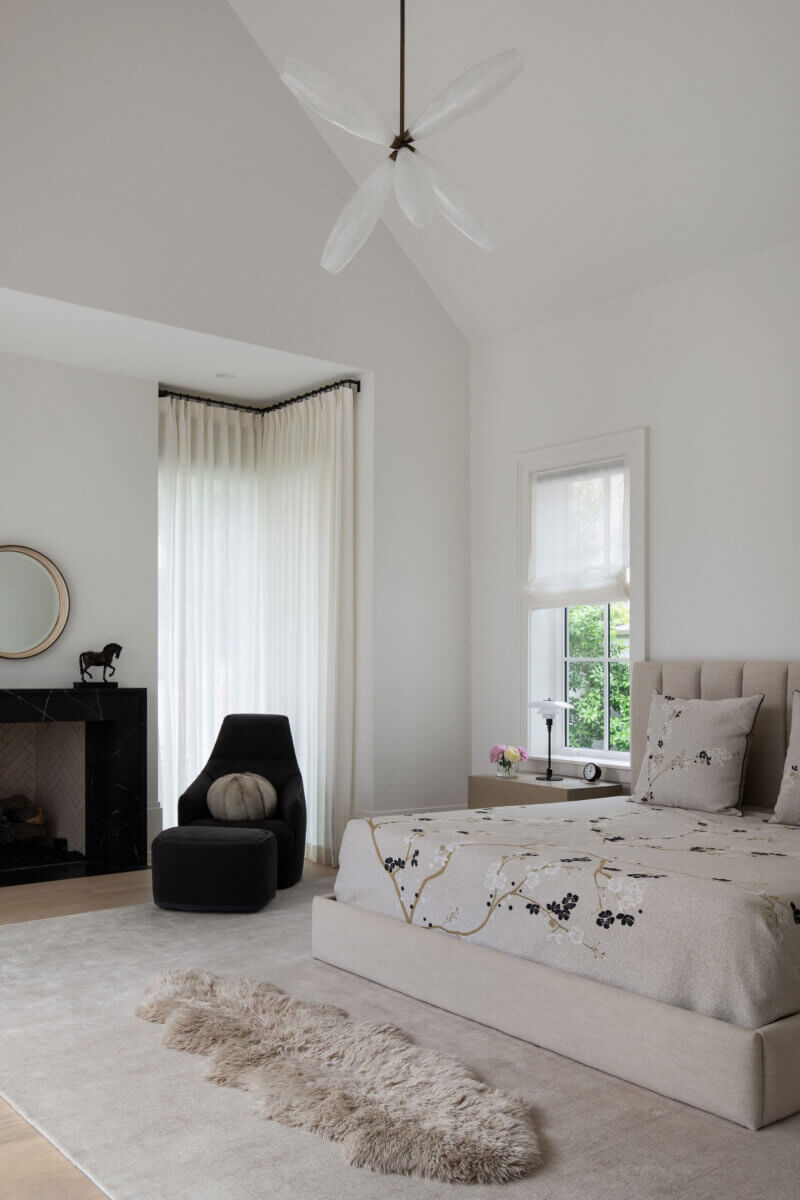 This Dallas Home Masters the Art of Quiet Luxury - 14 Modern Dallas home bedroom with beige bed, floral bedding, black armchair, fireplace, round mirror, fluffy rug, and large window with sheer white curtains—perfect for an NYC family ready to move south.