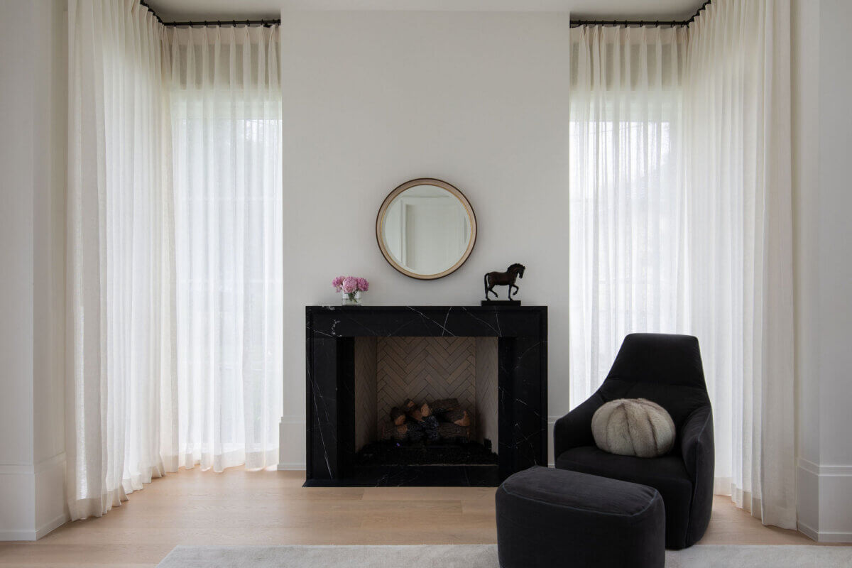 This Dallas Home Masters the Art of Quiet Luxury - 13 A modern Dallas home living room features sheer white curtains, a black marble fireplace with a round mirror and horse figurine on the mantel, plus a black armchair with a round pillow and ottoman.