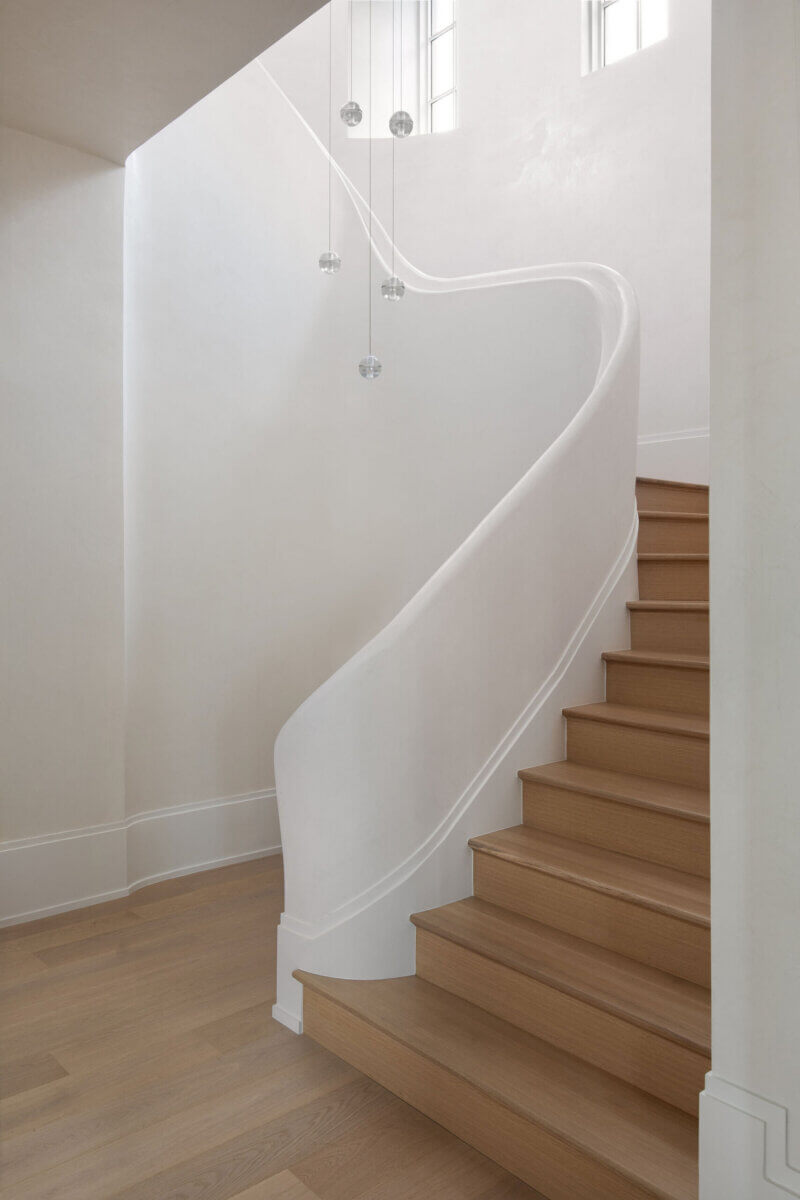 This Dallas Home Masters the Art of Quiet Luxury - 15 A modern wooden staircase with a white curved banister, light wood steps, and hanging glass pendant lights, illuminated by natural light from high windows.
