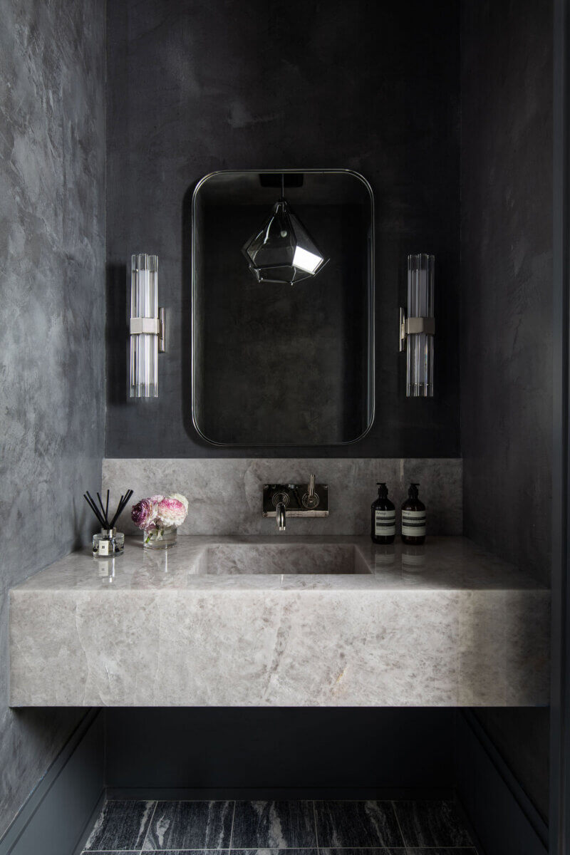 This Dallas Home Masters the Art of Quiet Luxury - 16 A modern bathroom in a modern Dallas home features dark gray walls, a rectangular marble sink, a large mirror, two wall sconces, and toiletries arranged neatly on the counter.