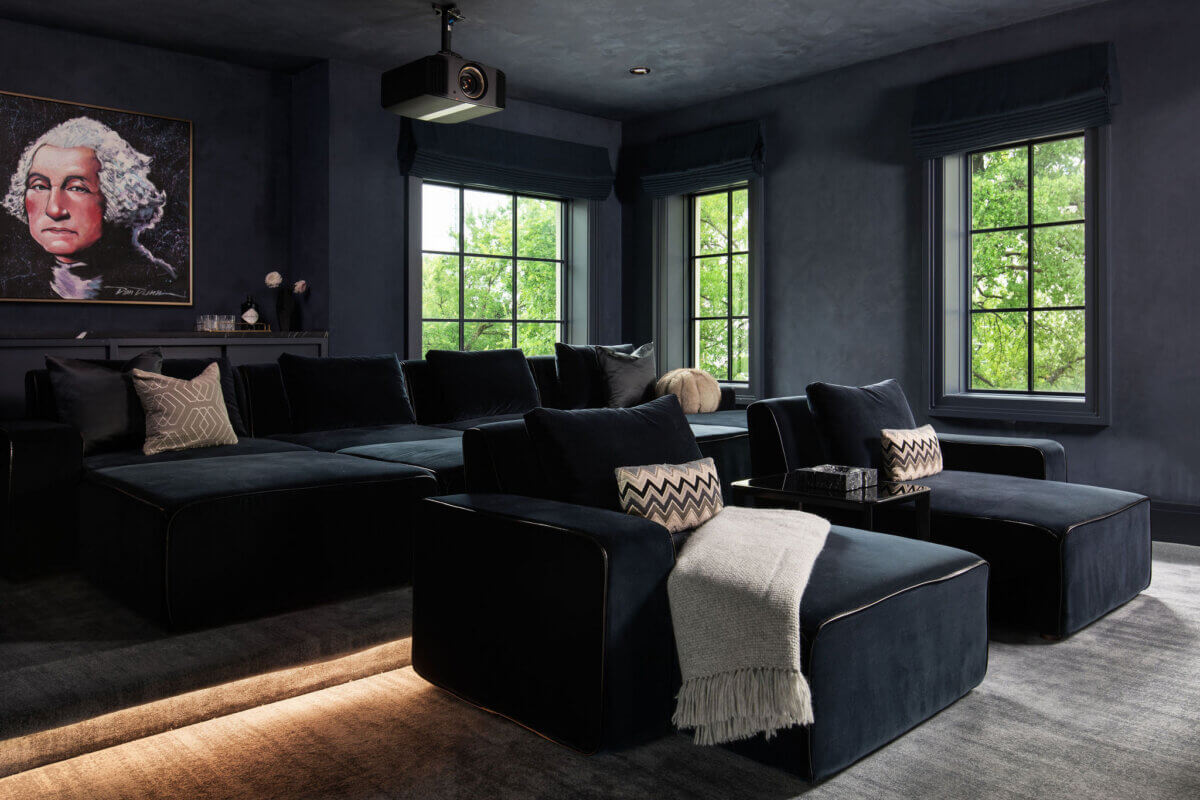 This Dallas Home Masters the Art of Quiet Luxury - 17 A modern Dallas home theater room with dark walls, large black seating, a projector, and three windows; a portrait of George Washington hangs on the wall.