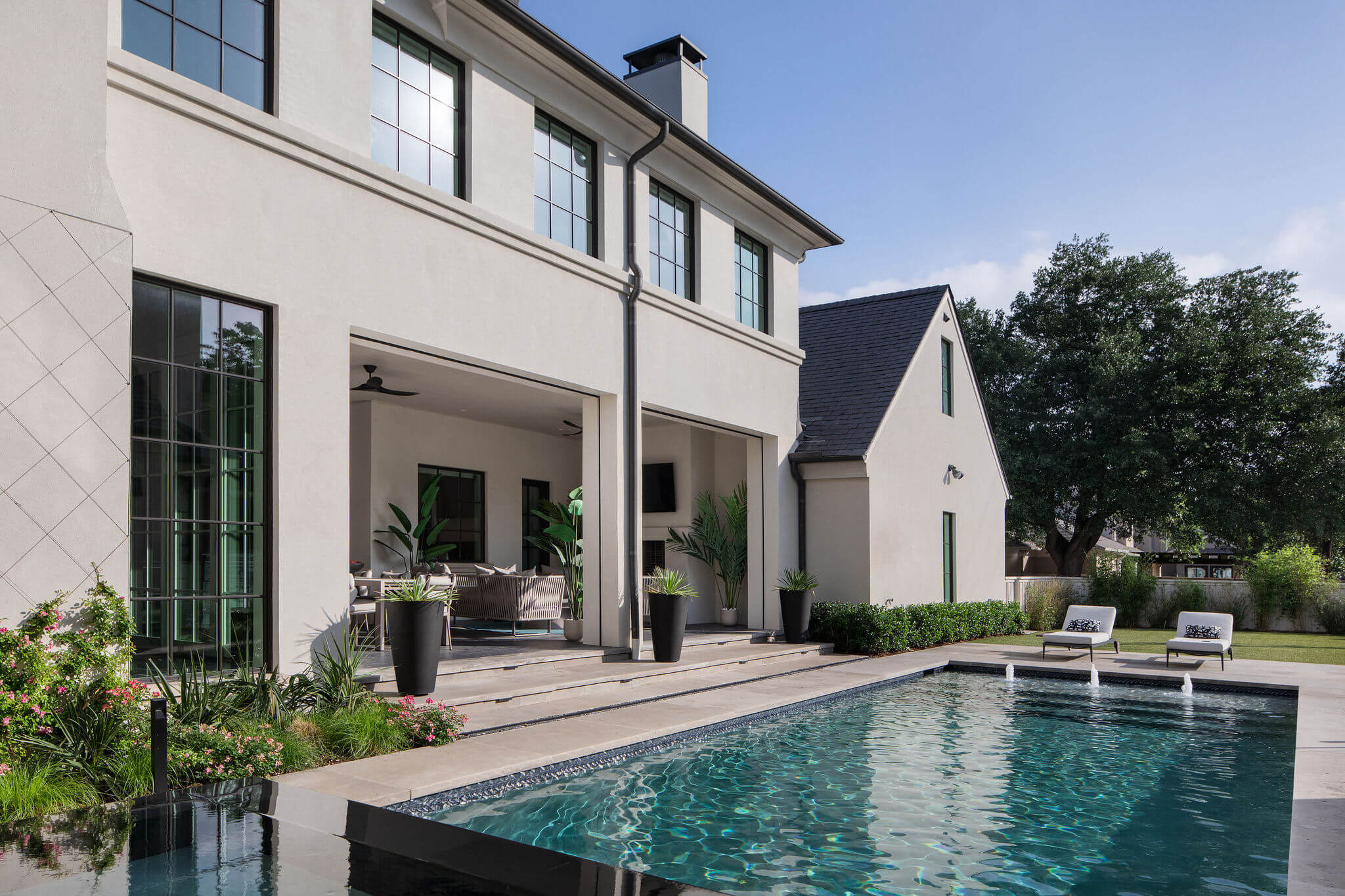 Modern Dallas home featuring large windows, an outdoor patio, potted plants, and a rectangular swimming pool with two lounge chairs beside it.