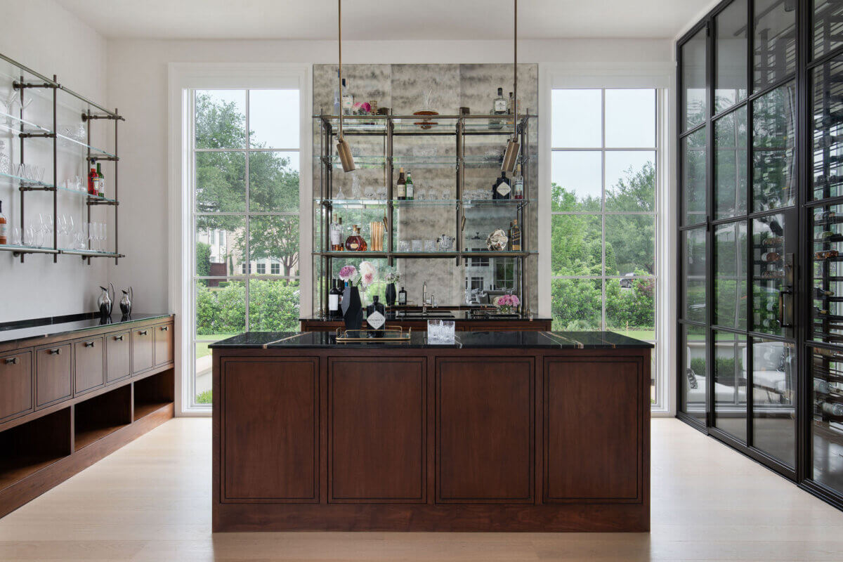 This Dallas Home Masters the Art of Quiet Luxury - 6 Modern Dallas home bar with a dark wood island, mirrored shelving with glassware, large windows, and glass doors leading to wine storage.