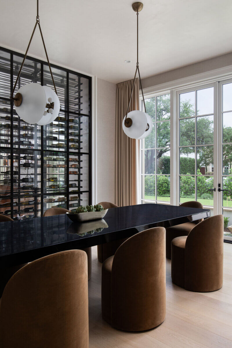 This Dallas Home Masters the Art of Quiet Luxury - 5 Modern dining room in a modern Dallas home featuring a black rectangular table, six brown upholstered chairs, two spherical pendant lights, large windows, and an indoor plant centerpiece.