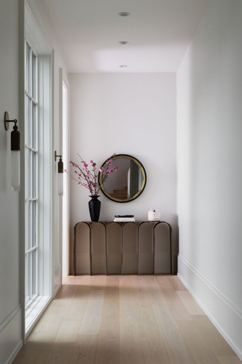 This Dallas Home Masters the Art of Quiet Luxury - 7 A narrow hallway in a modern Dallas home with light wood floors, a console table, round mirror, pink flowers, books, and a candle, all aglow with natural light from a glass door.