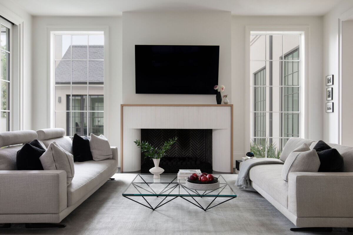 This Dallas Home Masters the Art of Quiet Luxury - 8 Modern living room in a modern Dallas home with two light gray sofas, a glass coffee table, white fireplace, wall-mounted TV, large windows, and minimal decor including a white vase and bowl of red apples.