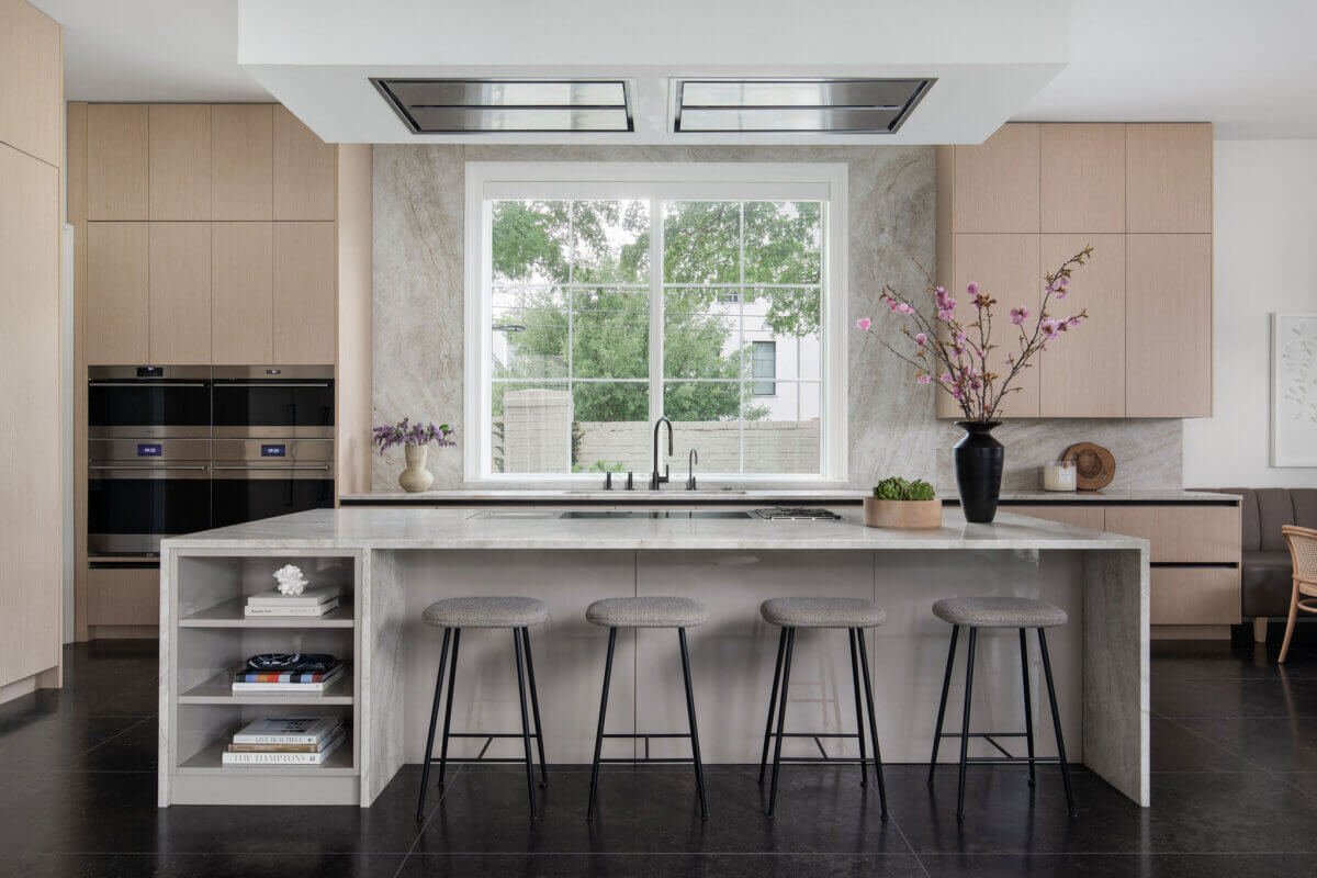 This Dallas Home Masters the Art of Quiet Luxury - 9 Modern kitchen in a Dallas modern home with a marble island, four gray barstools, built-in ovens, open shelving, large window, and decorative vases.