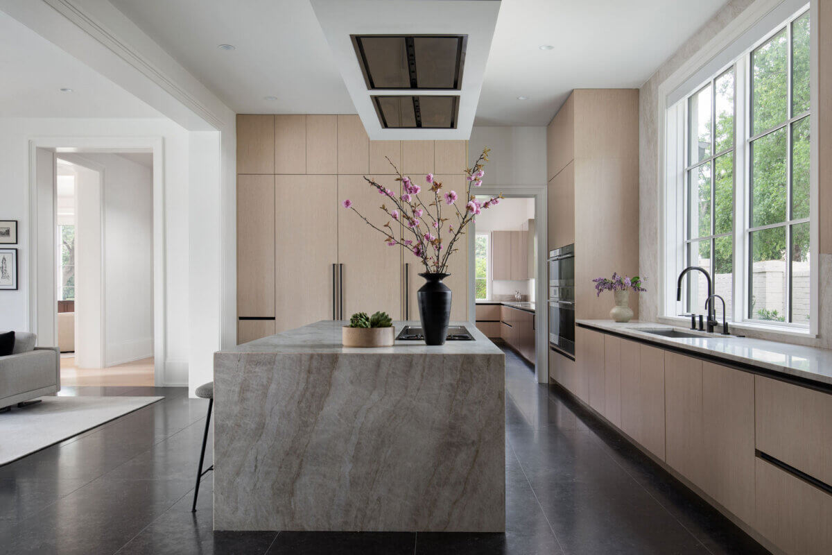 This Dallas Home Masters the Art of Quiet Luxury - 10 Minimalist modern kitchen with light wood cabinets, a marble island with floral arrangement, black countertops, large windows, and built-in stainless steel appliances—ideal for an NYC family seeking the comfort of a stylish modern Dallas home.