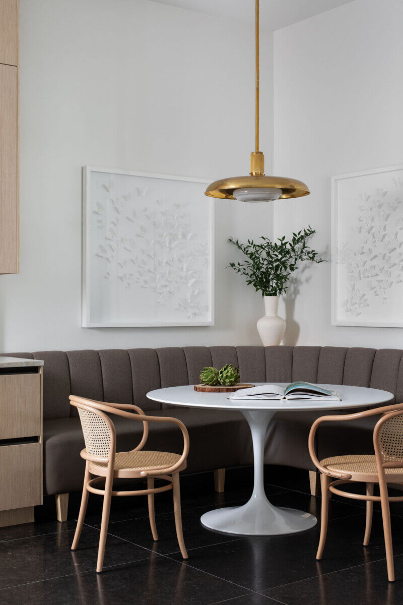 This Dallas Home Masters the Art of Quiet Luxury - 11 A modern Dallas home dining nook with a round white table, two wooden chairs, a curved bench, a gold pendant light, framed wall art, and a vase with greenery. An open book rests on the table.