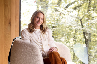 Mary Celeste Beall, proprietor of Blackberry Farm, sits smiling on a textured chair by a large window with greenery outside, her wavy blond hair complementing her white blouse and brown pants.