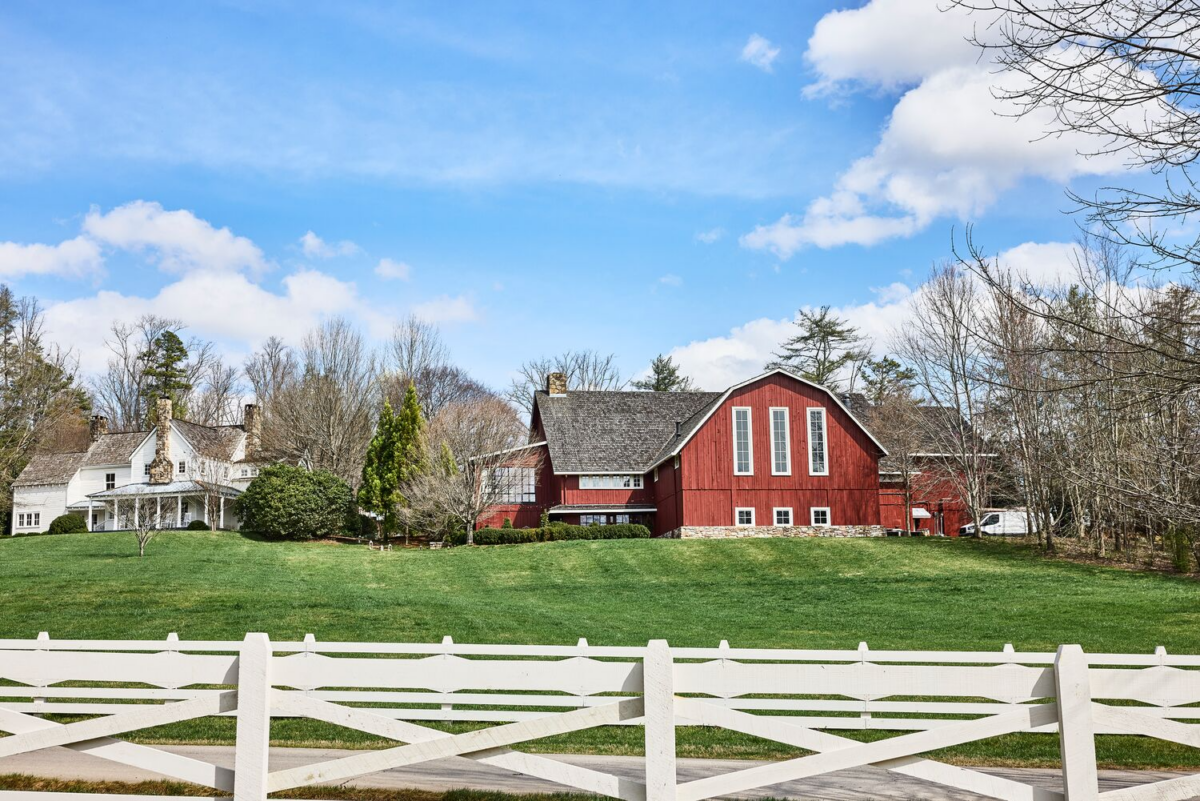 Meet the Woman Behind Tennessee's Beloved Blackberry Farm - 3 A large red barn and a white farmhouse sit on a grassy hill behind a white fence at Blackberry Farm, with trees and a blue sky in the background.
