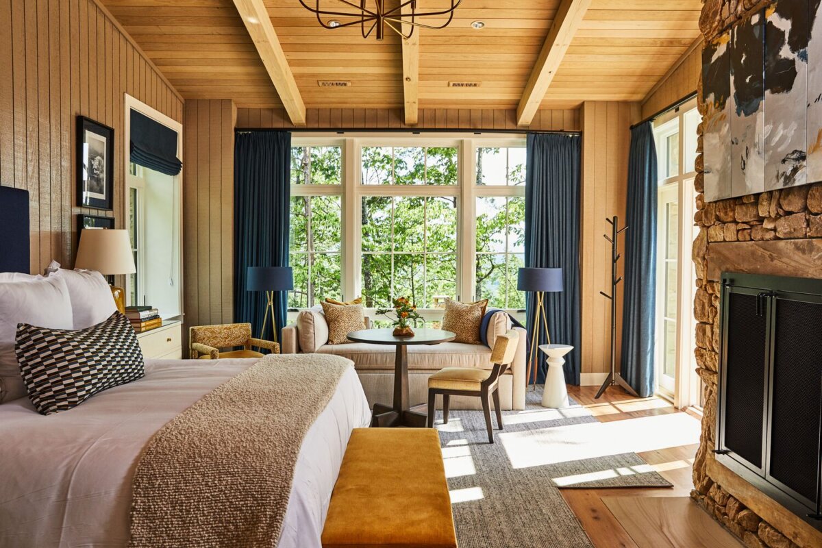Meet the Woman Behind Tennessee's Beloved Blackberry Farm - 4 A cozy bedroom at Blackberry Farm, featuring a large bed, sitting area, stone fireplace, and floor-to-ceiling windows with blue curtains overlooking greenery.