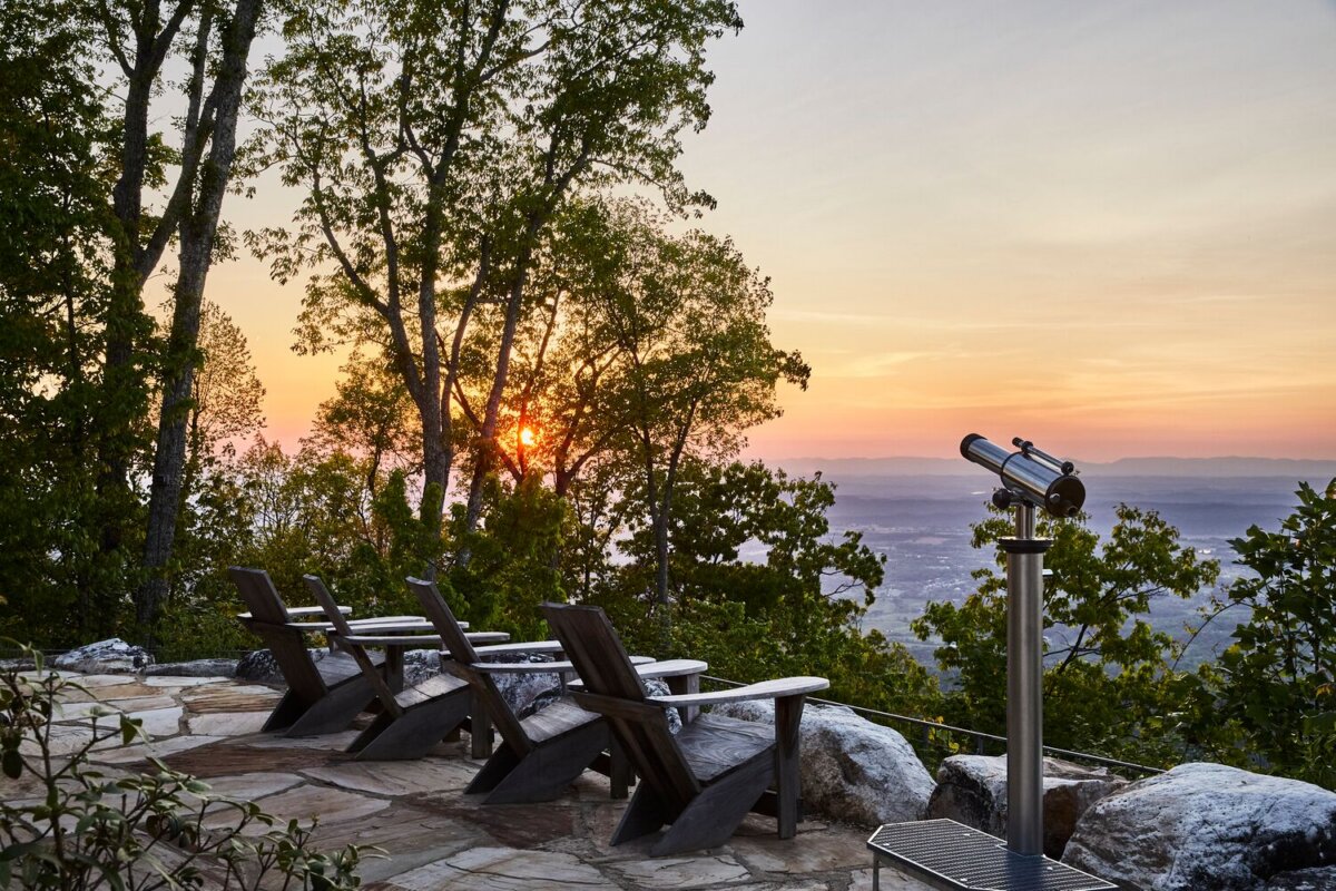 Meet the Woman Behind Tennessee's Beloved Blackberry Farm - 7 Several wooden chairs and a telescope are set on a stone patio overlooking a valley and forest at sunset at Blackberry Farm.