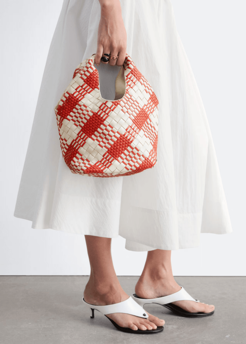 A person in a white skirt and white heeled sandals holds a red and white woven bag with a cut-out handle—perfect for showcasing colorful bags in style this season.