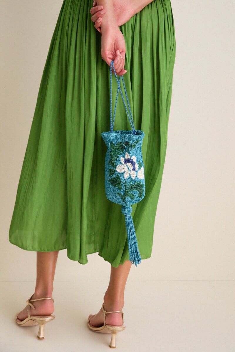 A person in a green skirt and heeled sandals holds one of this season's colorful bags—a blue beaded bag with a floral design and tassel—behind their back.
