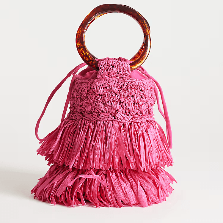 A pink raffia bucket bag with tiered fringe, a drawstring closure, and round brown handles—this season’s must-have for lovers of colorful bags—displayed upright against a neutral background.
