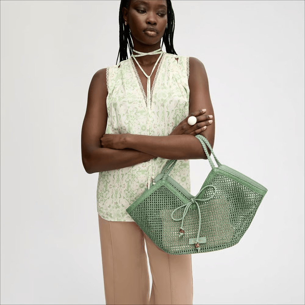 A woman stands with her arms crossed, wearing a light green patterned sleeveless top, beige pants, and holding one of this season’s must-have colorful bags—a large green woven handbag.