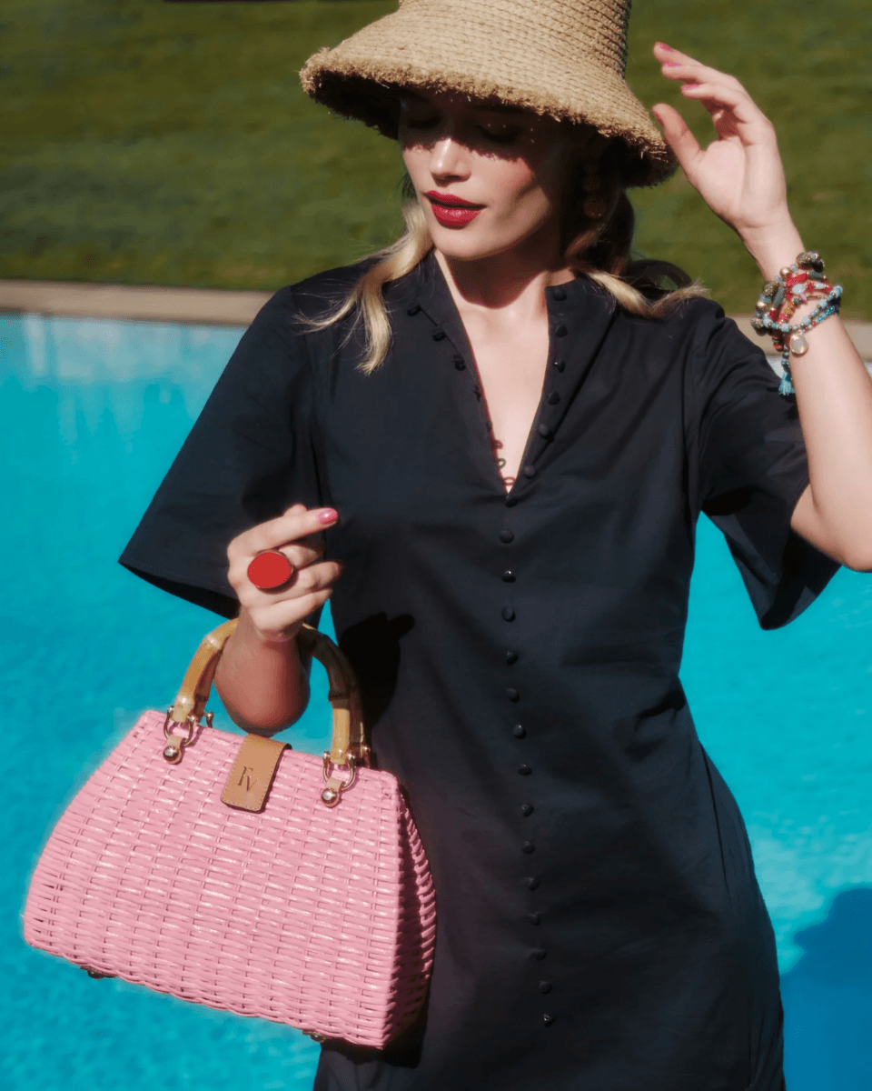 A woman in a black dress and straw hat stands by a pool, holding a pink woven handbag from this season's collection.