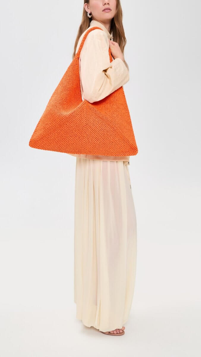 Woman wearing a long cream dress and light blouse, carrying one of this season’s colorful bags—an oversized orange woven triangular accessory—over her shoulder.