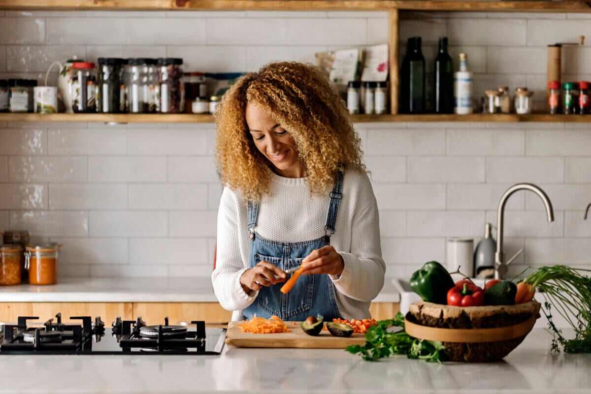 Person standing in a kitchen, smiling while slicing carrots on a wooden cutting board, surrounded by vegetables and kitchen items—preparing a meal that supports gut health and boosts metabolism.