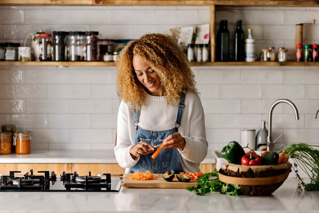 Person standing in a kitchen, smiling while slicing carrots on a wooden cutting board, surrounded by vegetables and kitchen items—preparing a meal that supports gut health and boosts metabolism.