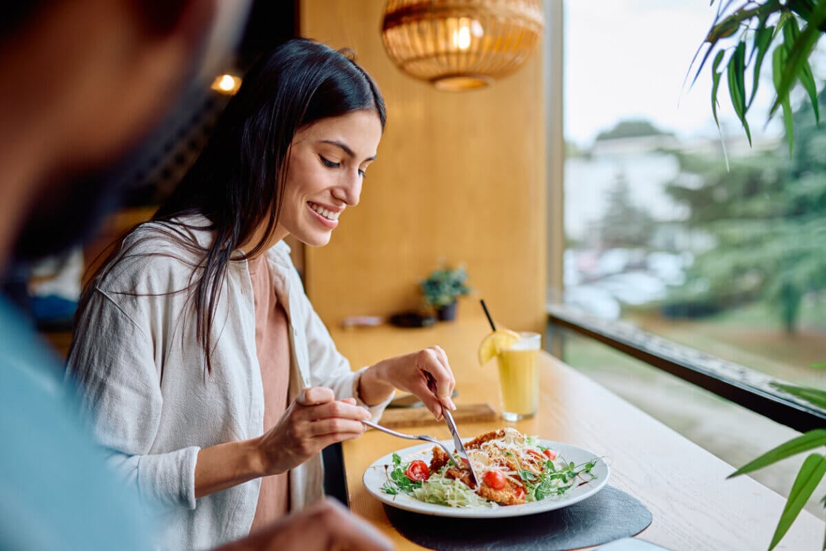 Your Gut Already Makes Its Own Ozempic — Here's How to Boost It! - 2 A woman sits by a window in a restaurant, smiling as she enjoys a meal with a fork and knife. A glass of lemonade is on the table next to her, reflecting her focus on gut health and mindful eating.